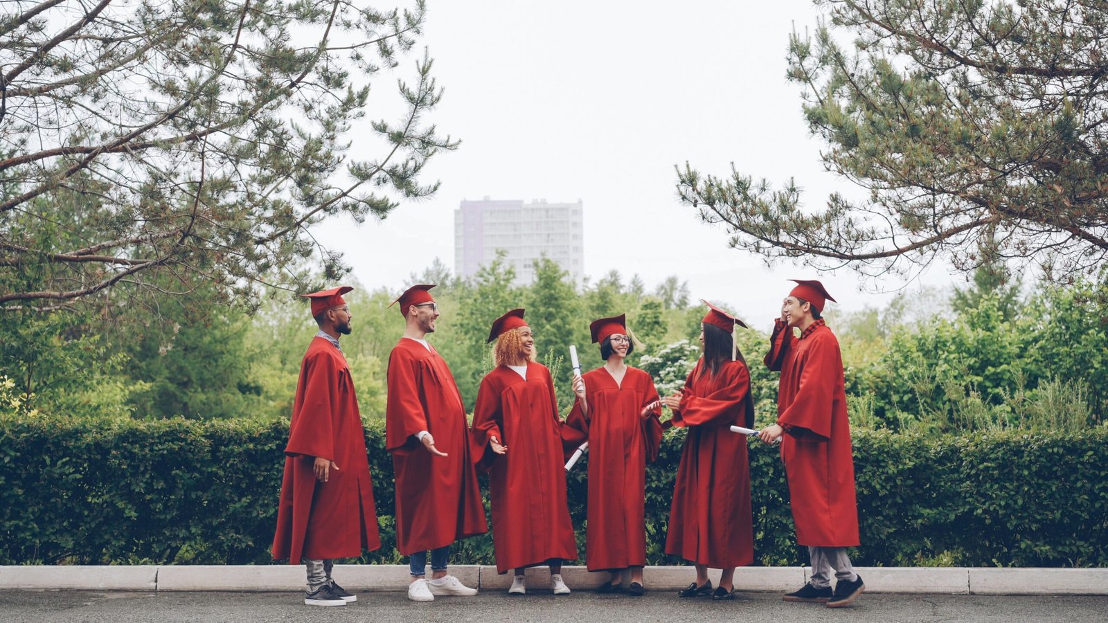 Graduates in red gowns stand together outdoors.